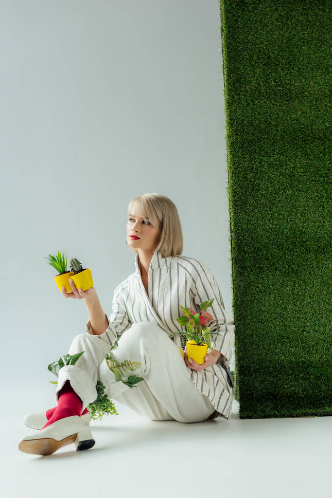 bella ragazza alla moda guardando lontano mentre tiene vasi di fiori sul grigio con erba verde
  - Foto, immagini