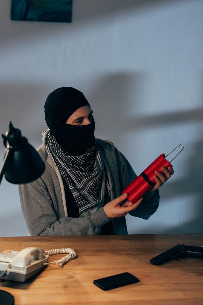 Terrorist in mask holding dynamite while sitting in dark room - Photo, Image