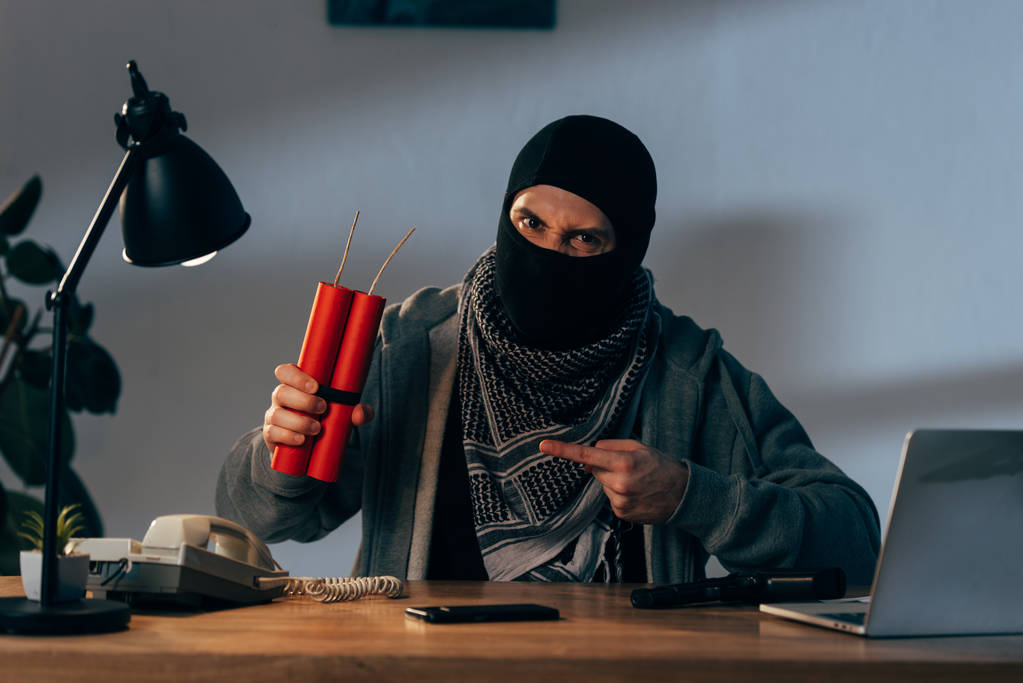 Terrorista enojado en máscara sentado en la mesa y señalando con el dedo a la dinamita
 - Foto, imagen