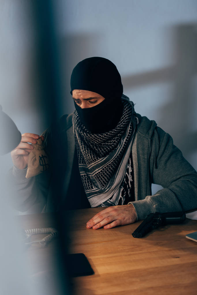 Molesto criminal con máscara negra sentado en la mesa y sosteniendo una pequeña bolsa de dinero
 - Foto, imagen