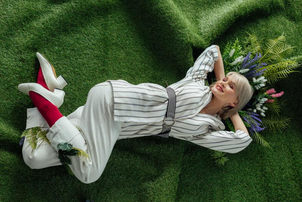 high angle view of beautiful stylish girl smiling while lying on artificial grass with flowers  - Photo, Image