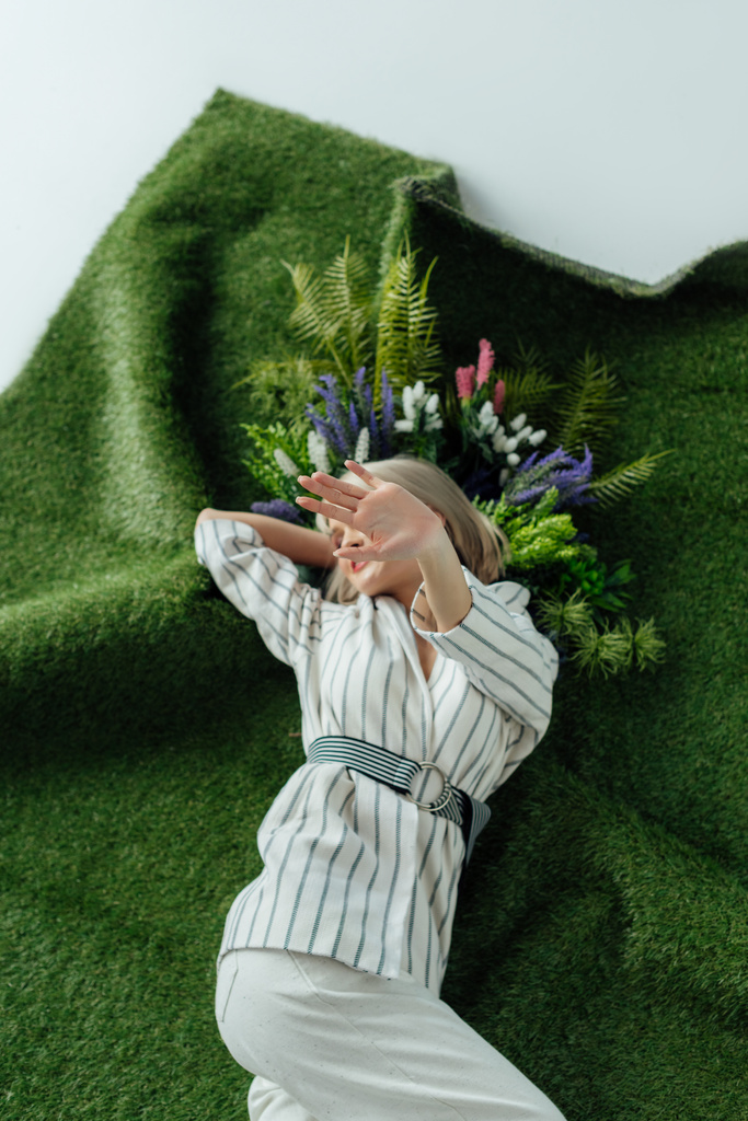 シダと花と人工芝生の上に横たわっている間手で顔を覆っているスタイリッシュな女の子 ロイヤリティフリー写真 画像素材