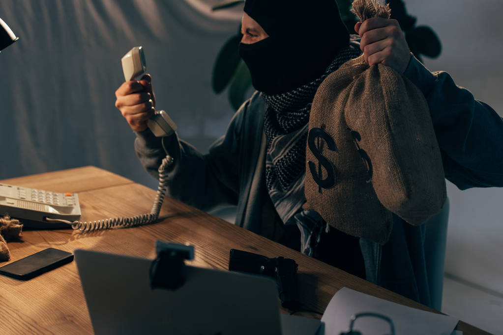 Terrorista enojado con máscara negra sosteniendo bolsas de dinero y mirando el auricular
 - Foto, Imagen