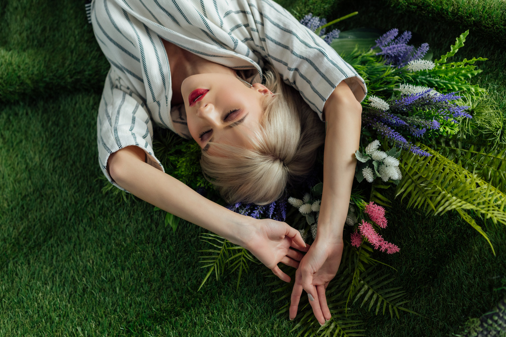 vista de ángulo alto de hermosa chica elegante con los ojos cerrados acostado en césped artificial con helecho y flores
 - Foto, Imagen