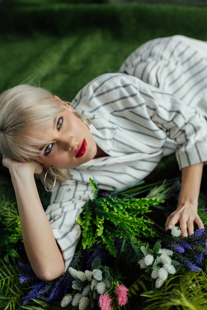mooi stijlvol meisje camera kijken terwijl liggend op kunstgras met varens en bloemen - Foto, afbeelding