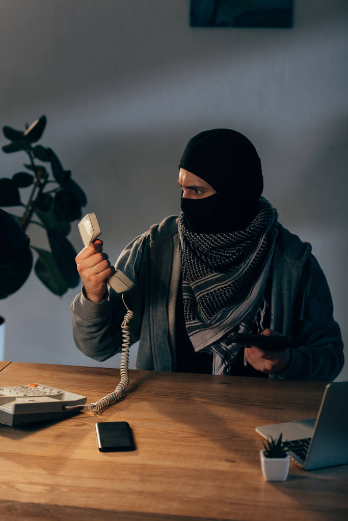 Terorista v černé masce, drží zbraň a při pohledu na sluchátko - Fotografie, Obrázek