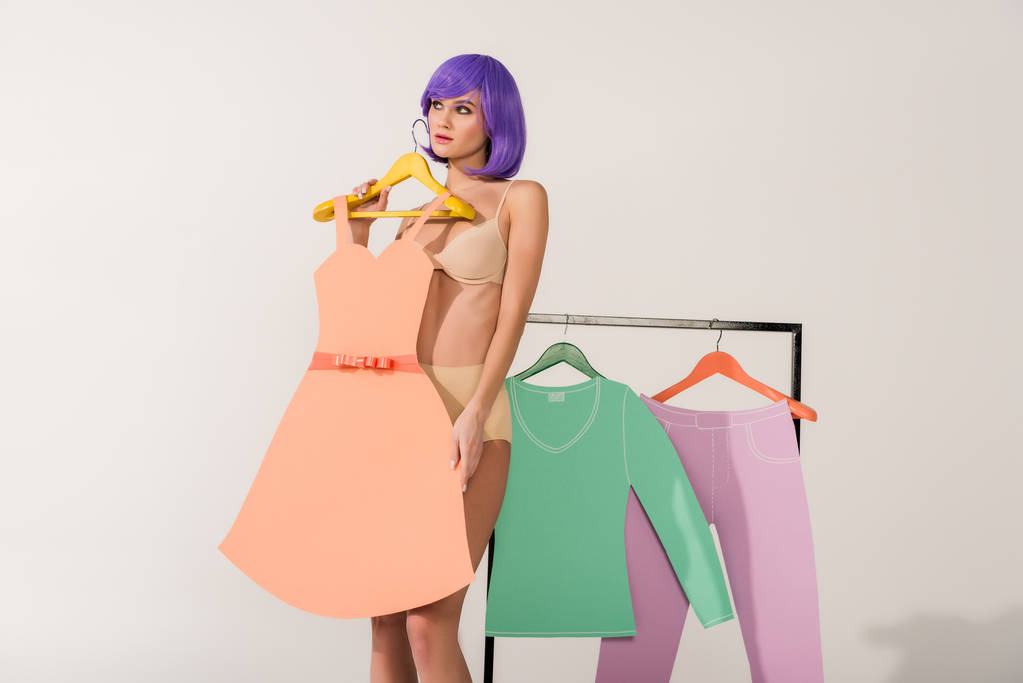 chica atractiva con pelo morado sosteniendo vestido de papel y posando cerca de rack con ropa de colores en blanco
  - Foto, imagen