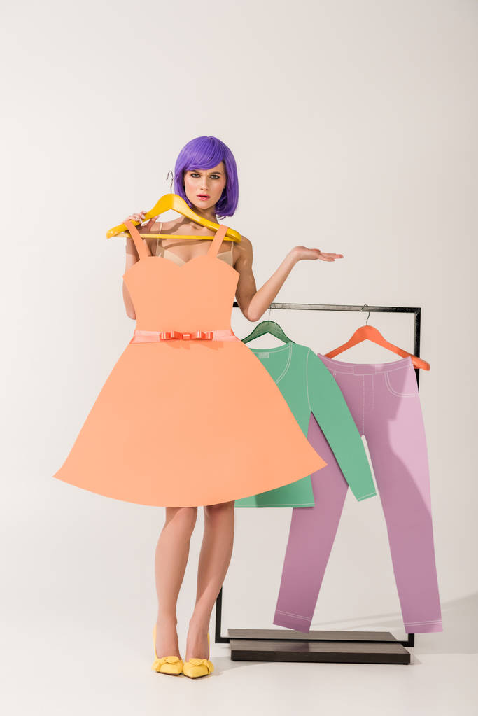 beautiful surprised girl with purple hair holding paper dress and posing near rack with colorful clothes on white  - Photo, Image