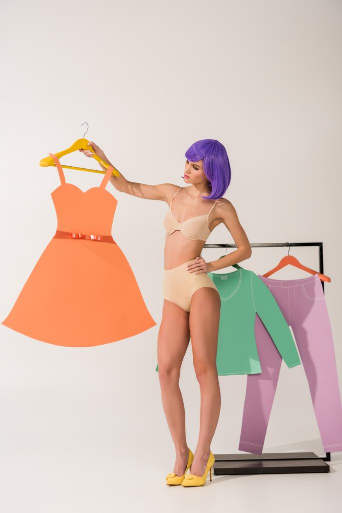 beautiful girl with purple hair in underwear holding paper dress and posing near rack with colorful clothes on white  - Photo, Image
