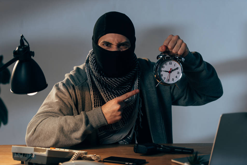 Злой террорист в черной маске указывает пальцем на будильник
 - Фото, изображение