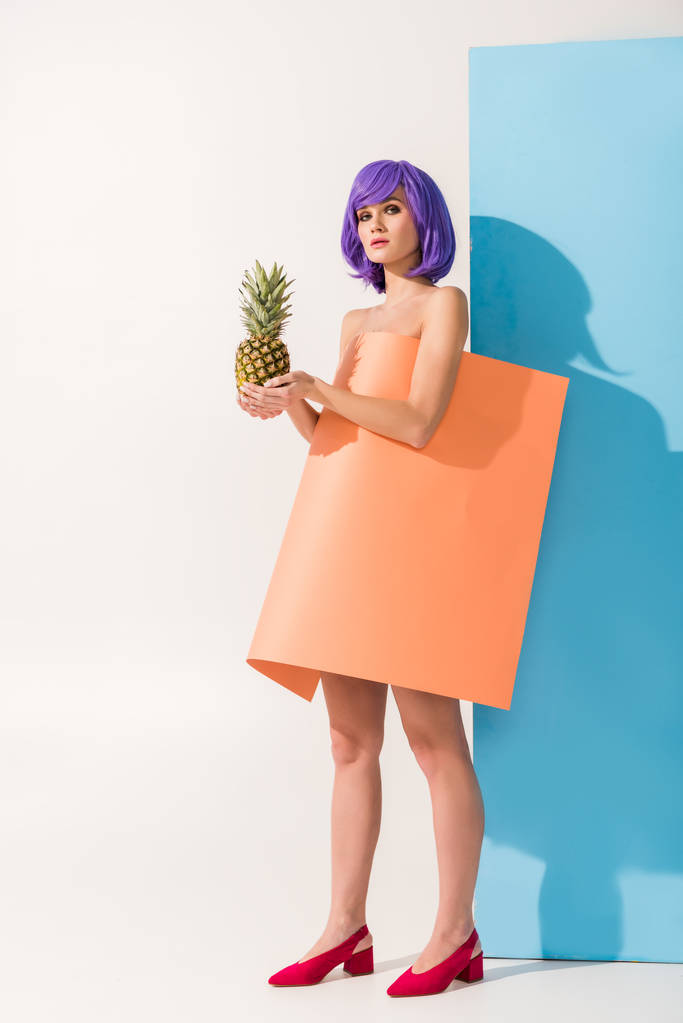 beautiful girl with purple hair covered in coral paper sheet holding pineapple while looking at camera on blue and white - Photo, Image