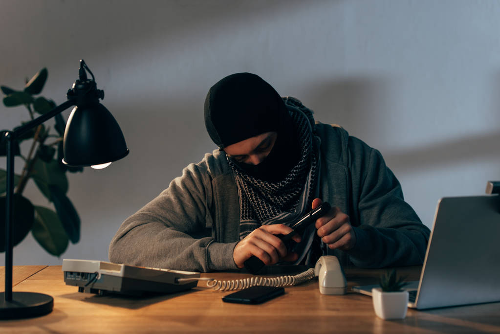 Terrorist in zwart masker zittend aan tafel ingedrukt te houden pistool - Foto, afbeelding