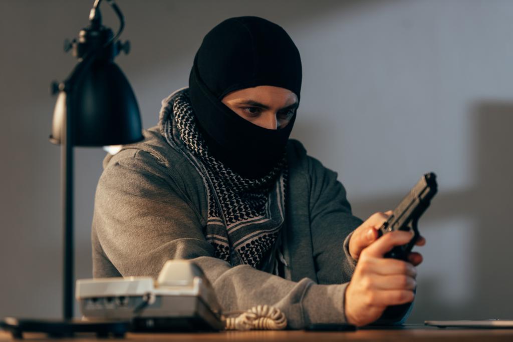 Concentrated criminal in black mask loading gun in room - Photo, Image