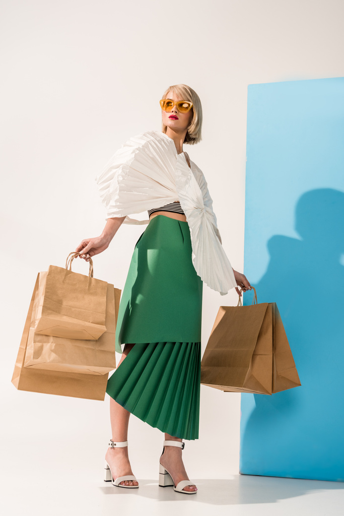 kaunis tyylikäs nuori nainen aurinkolasit ja paperi vaatteet poseeraa ostoskassit sininen ja valkoinen
 - Valokuva, kuva