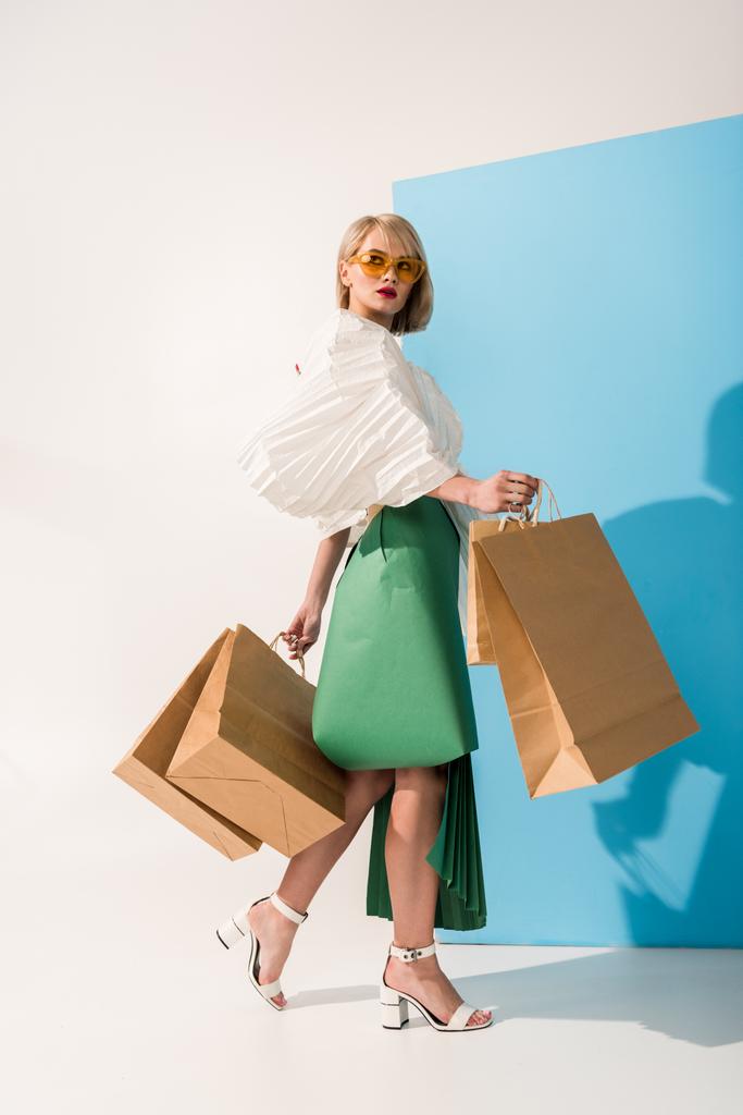 schöne stylische Mädchen in Sonnenbrille und Papierkleidung posiert mit Einkaufstaschen auf blau-weiß - Foto, Bild