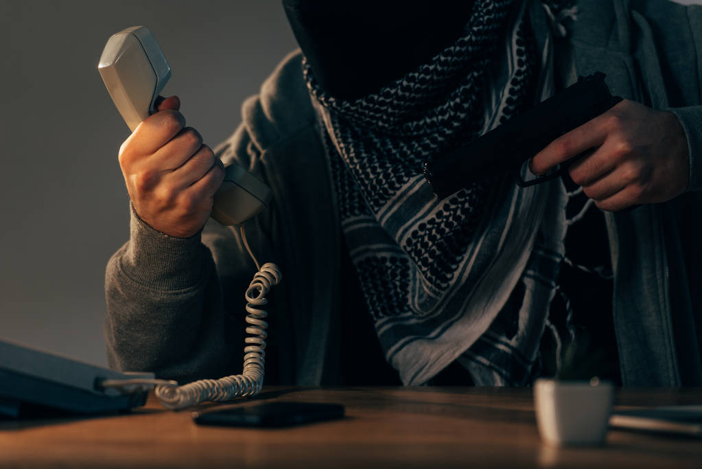 Обрізаний вид терориста, який тримає кишеньковий пристрій і пістолет за столом
 - Фото, зображення