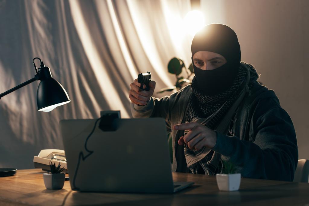 Verbrecher in Maske zeigt mit Finger und zielt mit Waffe auf Laptop - Foto, Bild