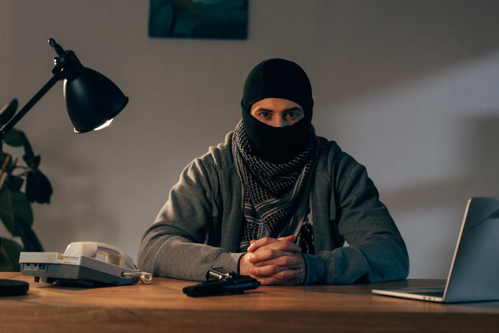 Terrorist in masker zit aan tafel met geïnterlinieerde vingers - Foto, afbeelding