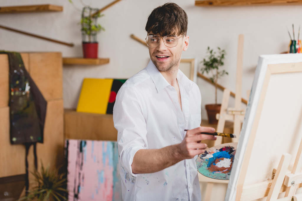 selective focus of smiling artist in white shirt and glasses painting on canvas - Photo, Image