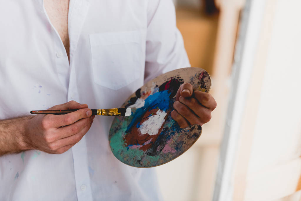 vista parcial del artista en camisa blanca mezclando pinturas con pincel en paleta
 - Foto, Imagen