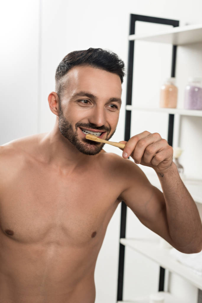 schöner hemdloser Mann beim Zähneputzen, während er im Badezimmer in den Spiegel schaut  - Foto, Bild