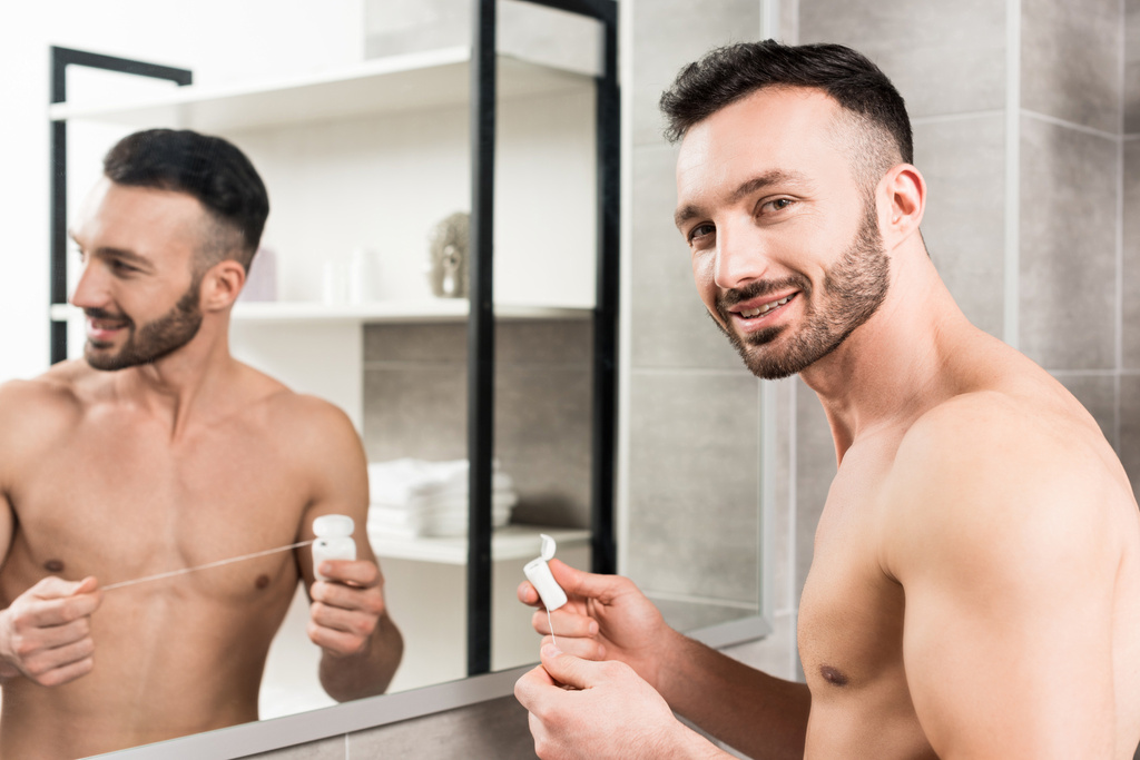 χαρούμενα γενειοφόρο άτομο που κρατά το οδοντικό νήμα ενώ στέκεται κοντά καθρέφτη στο μπάνιο  - Φωτογραφία, εικόνα