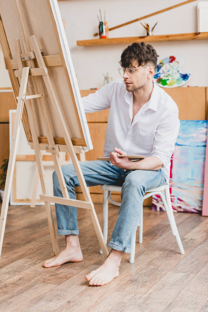 knappe kunstenaar in wit shirt en glazen schilderij op doek zittend op een stoel in schilderij studio - Foto, afbeelding