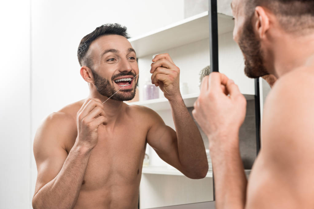 ευτυχισμένος άνθρωπος shirtless κοιτάζοντας καθρέφτη ενώ χρησιμοποιείτε οδοντικό νήμα στο μπάνιο  - Φωτογραφία, εικόνα