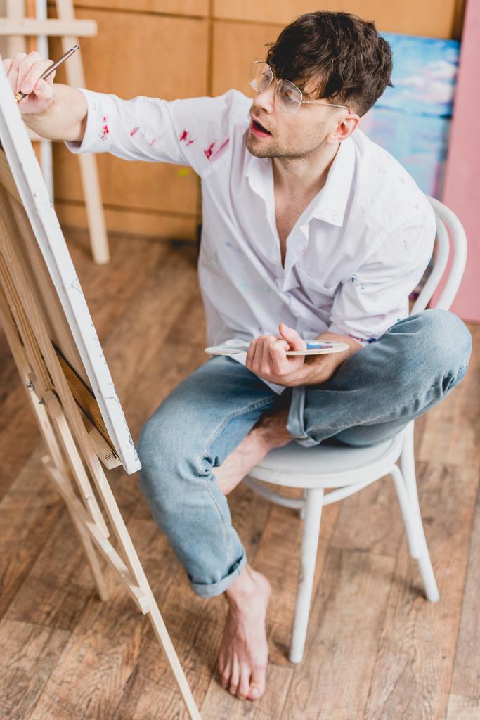 selektiver Fokus des gut aussehenden Künstlers auf Leinwand, während er auf einem Stuhl im Malatelier sitzt - Foto, Bild