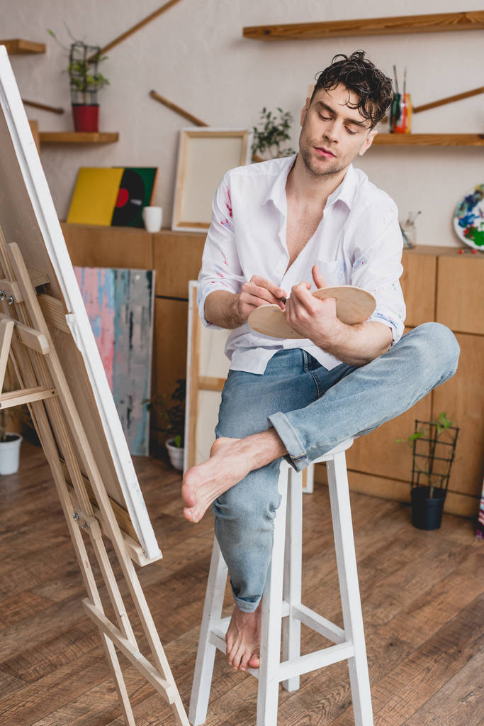 pohledný umělec v bílou košili a modré džíny sedí u stojanu v malířském ateliéru - Fotografie, Obrázek