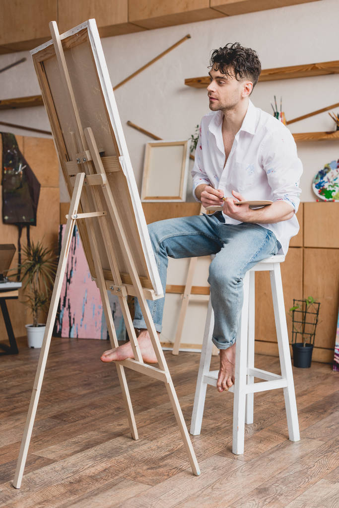 artista guapo en camisa blanca y vaqueros azules sentado en el caballete en el estudio de pintura
 - Foto, Imagen
