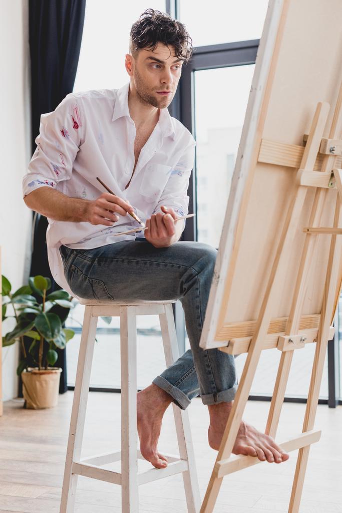 artista guapo en camisa blanca y vaqueros azules mezclando pinturas en la paleta mientras está sentado en el caballete en la galería
 - Foto, imagen