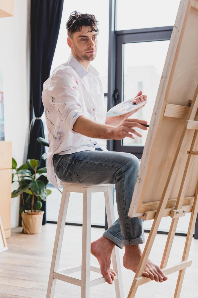 handsome artist in white shirt and blue jeans painting on canvas in gallery - Photo, Image