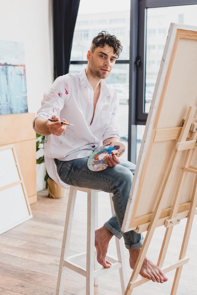 Beautiful artist with palette and paintbrush sitting on high chair and looking at camera
 - Фото, изображение