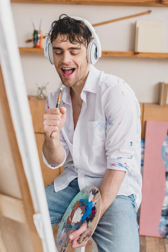 selective focus cheerful artist in headphones, singing with paintbrush while holding palette - Photo, Image