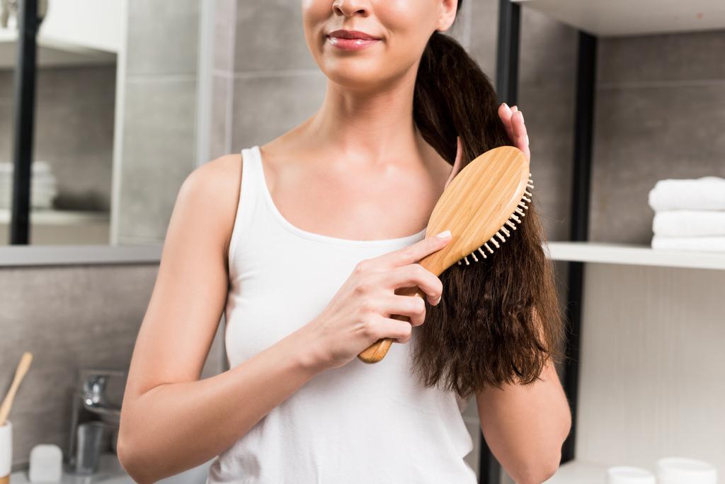 περικοπεί άποψη της χαμογελώντας μελαχρινή γυναίκα βούρτσισμα τα μαλλιά ενώ στέκεται στο μπάνιο  - Φωτογραφία, εικόνα