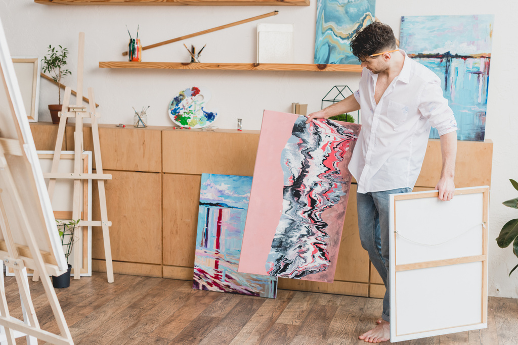 bosý umělec v bílou košili a modré džíny přenášení obrazů v galerii - Fotografie, Obrázek
