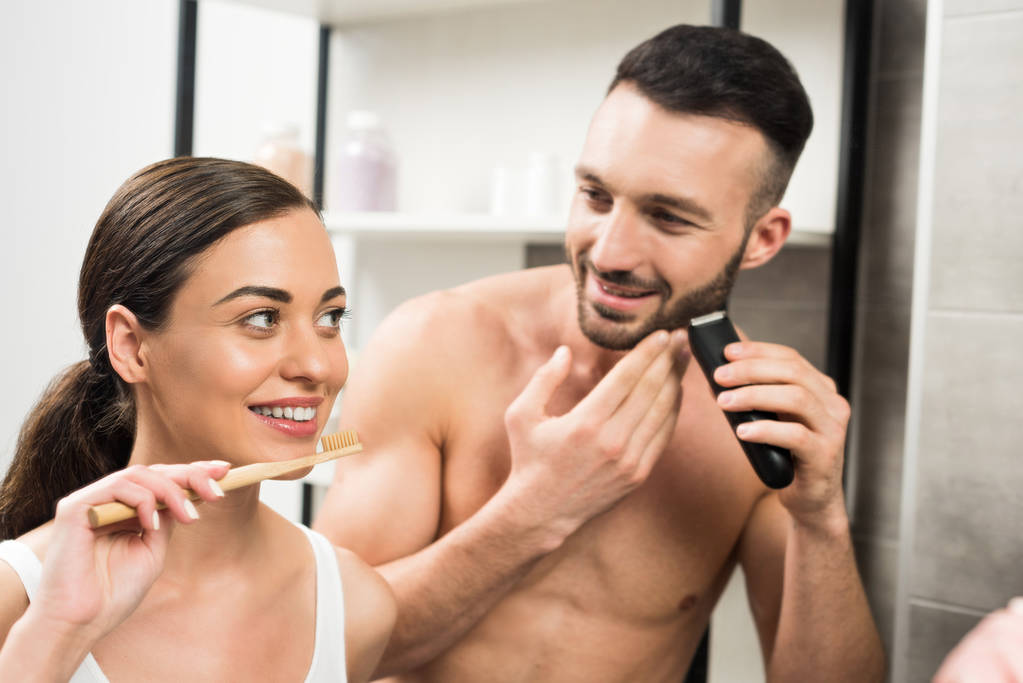 atractiva mujer sosteniendo cepillo de dientes cerca guapo sin camisa novio afeitándose la cara en el baño
  - Foto, imagen