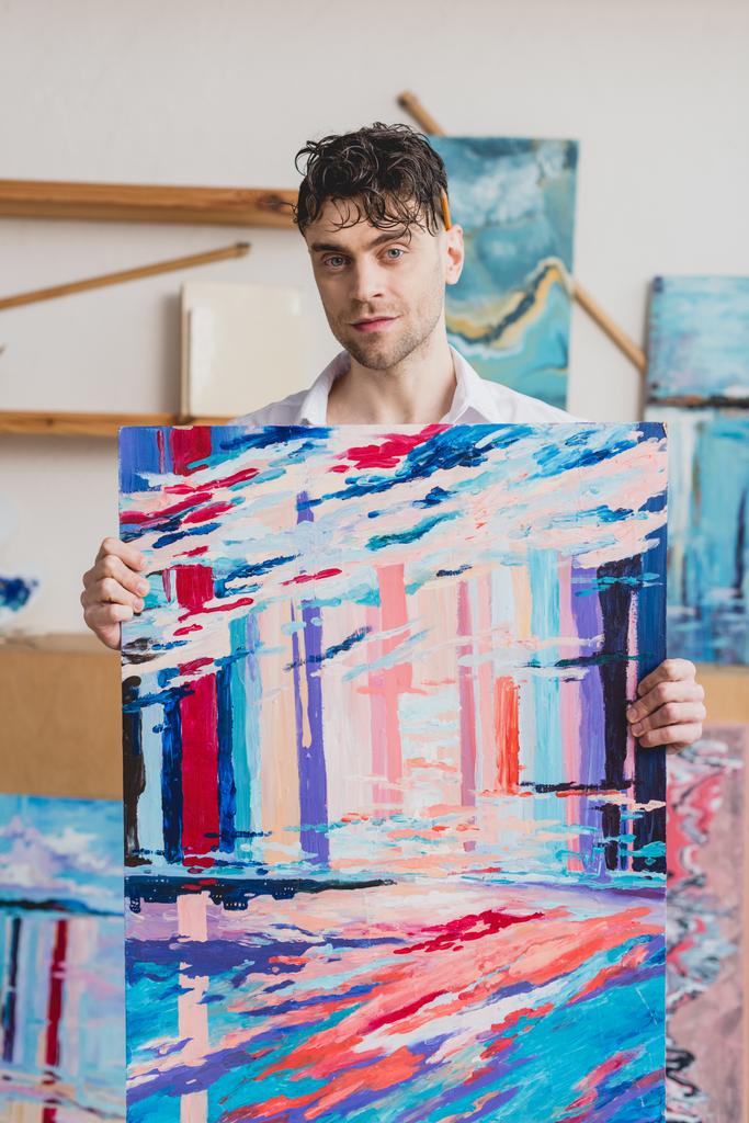 knappe kunstenaar holding veelkleurige abstract schilderen en camera kijken - Foto, afbeelding