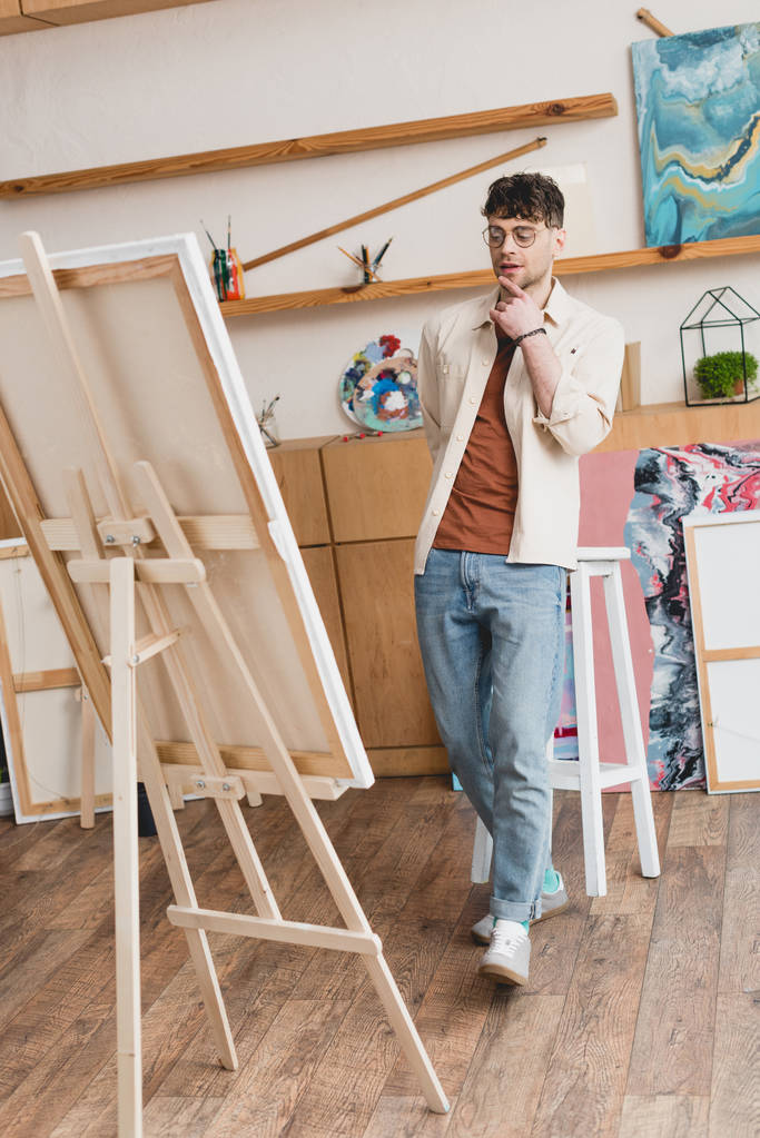 bel artiste en jeans bleus et chemise rose debout dans l'atelier de peinture et regardant chevalet avec toile
 - Photo, image