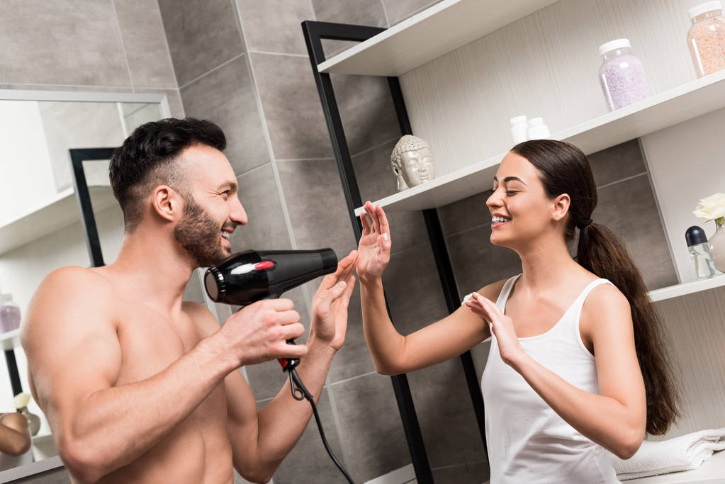 παιχνιδιάρικο γενειοφόρο άτομο που κρατά τα μαλλιά στεγνωτήρα ενώ στέκεται με χαρούμενα γυναίκα στο μπάνιο  - Φωτογραφία, εικόνα