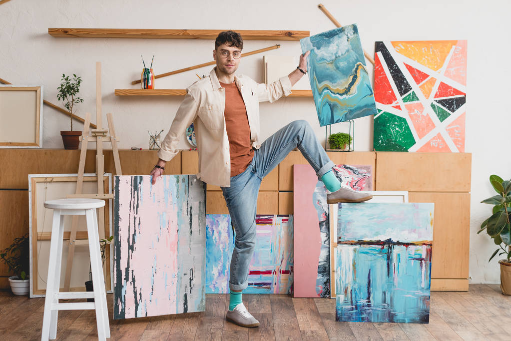 handsome artist in pink shirt and blue jeans standing in painting studio with multicolored abstract paintings - Photo, Image