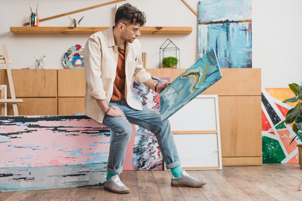 красивый художник в розовой рубашке и синих джинсах смотрит на красочную абстрактную живопись
 - Фото, изображение