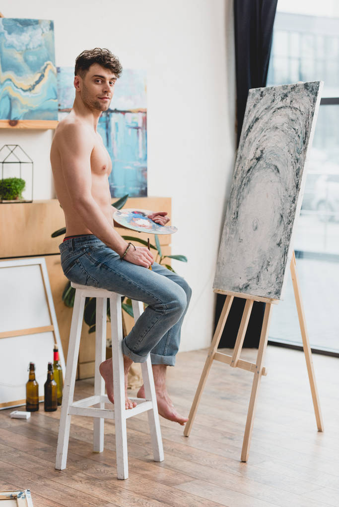 hyvännäköinen puolialaston taiteilija katselee kameraa istuessaan syöttötuolilla lähellä maalausta
 - Valokuva, kuva