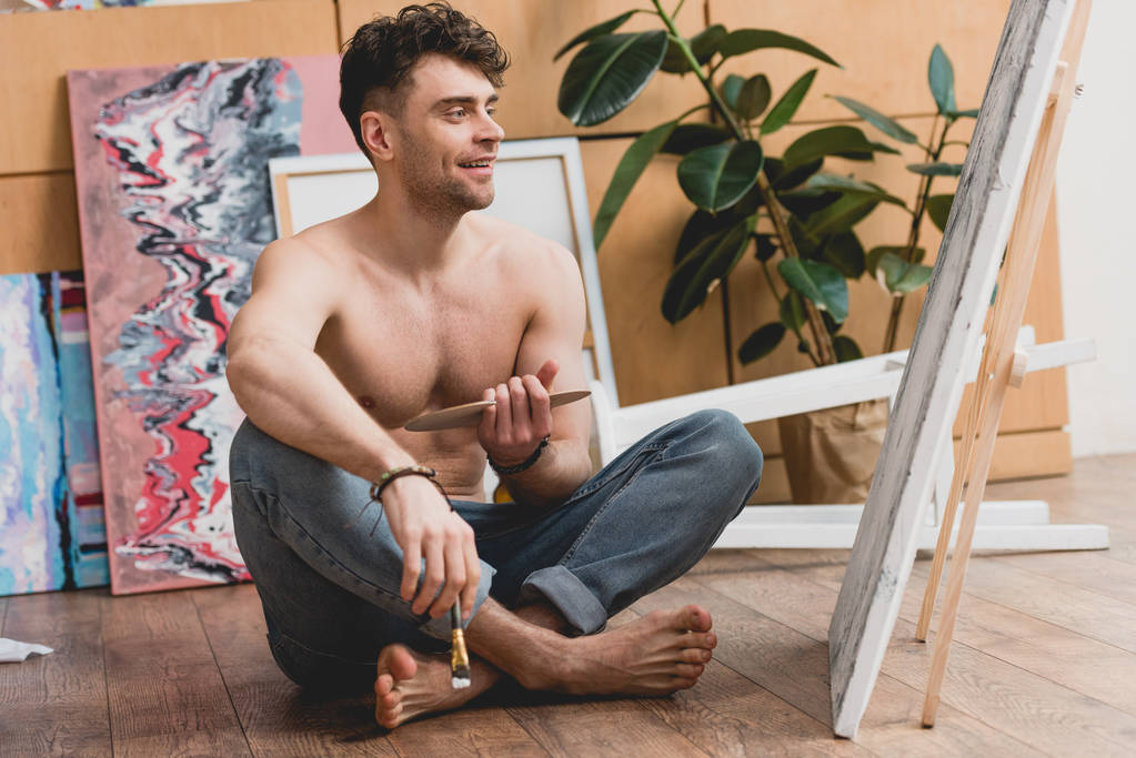 uśmiechający się artysta pół naga siedzi ze skrzyżowanymi nogami na podłodze w studio malarstwa - Zdjęcie, obraz