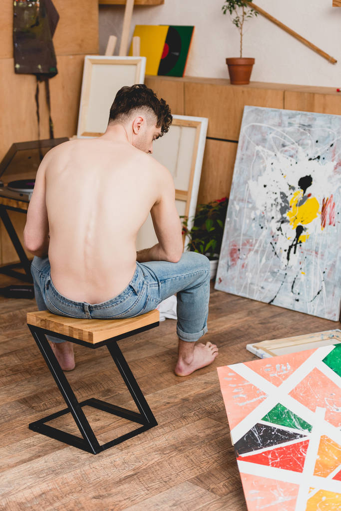 полуголый художник в синих джинсах сидит на стуле в галерее, окруженный картинами
 - Фото, изображение