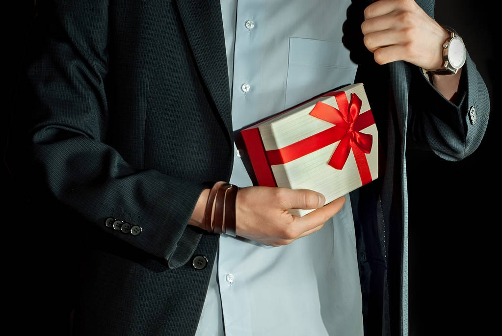 Ένας άνθρωπος σε ένα κοστούμι κατέχει ένα κουτί δώρου στα χέρια του. Πλαίσιο με μια κόκκινη κορδέλα και φιόγκο. - Φωτογραφία, εικόνα