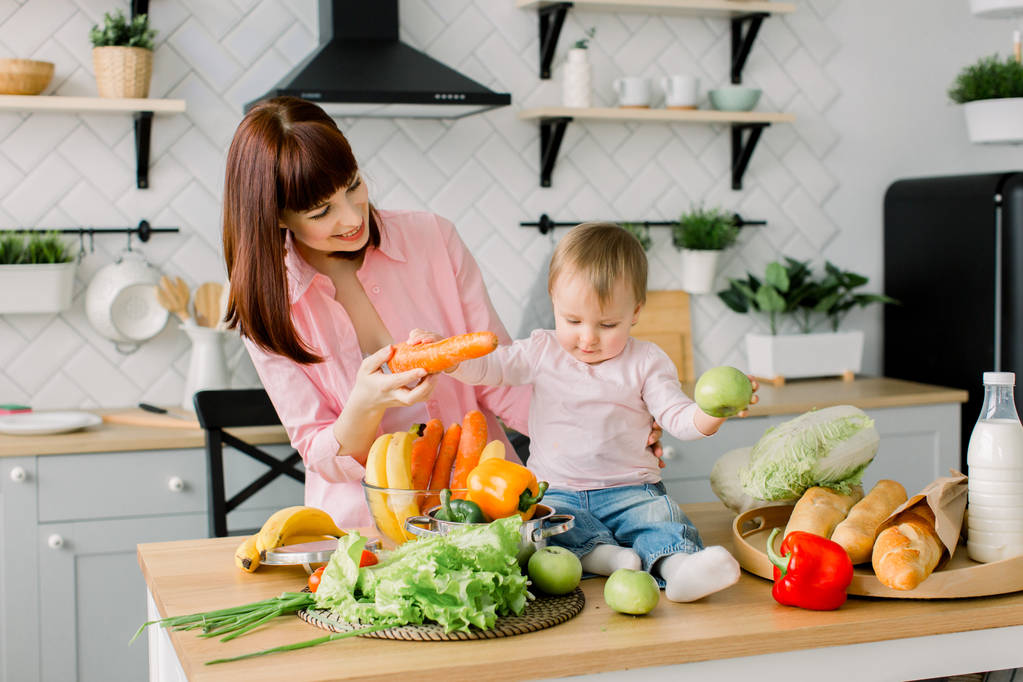 Onnellinen äiti esittelee tuoreita vihanneksia pienelle tyttövauvalleen, joka istuu puupöydällä kotikeittiössä. Perhe, ruoka, terveellinen ruokavalio, ruoanlaitto ja ihmisten käsite
 - Valokuva, kuva