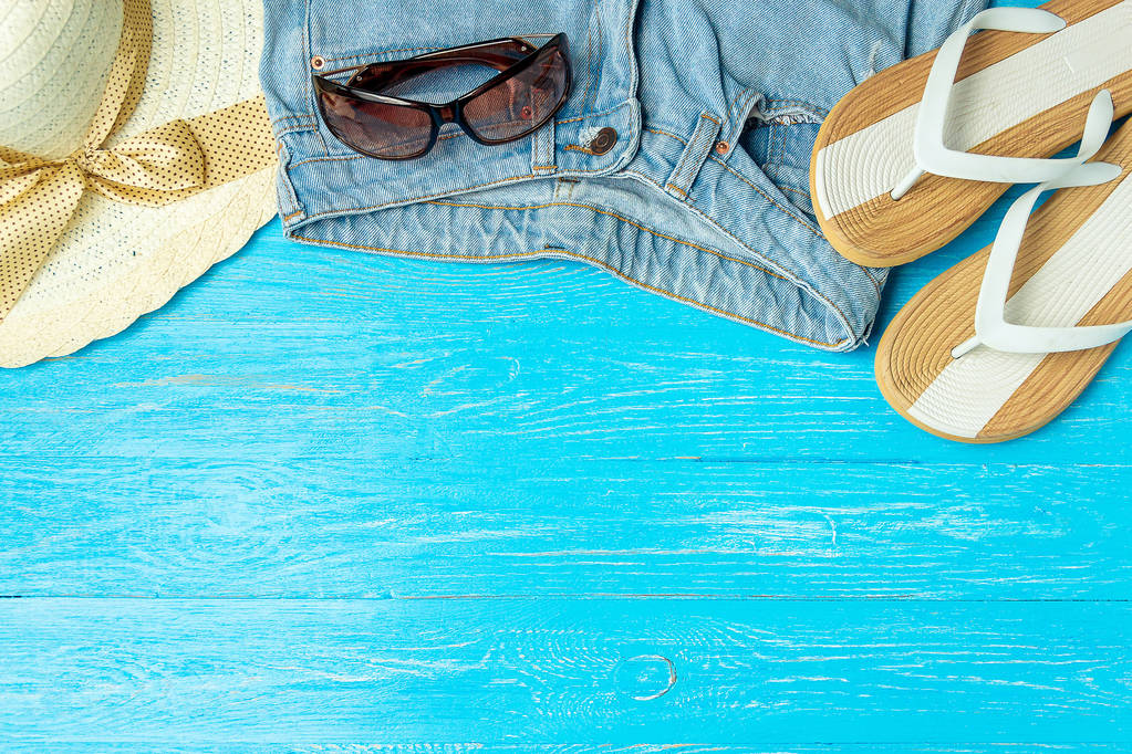 Πλαίσιο θηλυκό και κομψό καπέλο αχύρου παντόφλες τζιν γυαλιά ηλίου σε μπλε φόντο ξύλινη, copyspace για το κείμενο, καλοκαιρινές διακοπές. - Φωτογραφία, εικόνα