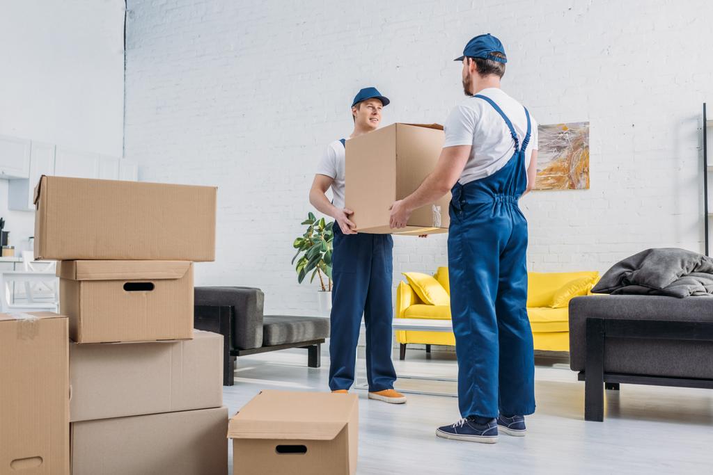dva stěhovací společnosti v uniformě přepravující kartonové krabice v obývacím pokoji - Fotografie, Obrázek